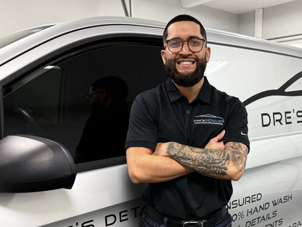 Andre Hernandez standing with his arms crossed beside his white Dre’s Detailing van, wearing a black shirt with the company logo.