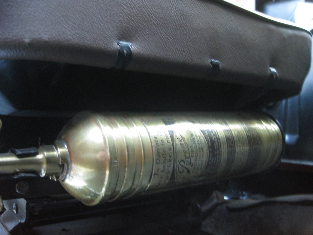A polished brass fire extinguisher with an engraved label, mounted beneath the leather edge of the driver's seat.