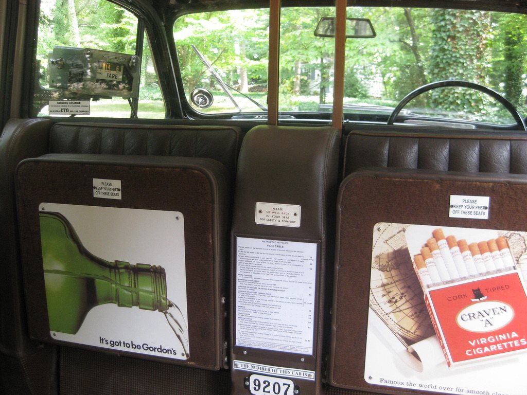 Looking forward from the rear seat: two jump seats folded up against the partition, each showing a period advertisement, with the printed fare table between them above a plate reading 9207, and the driver's seat and windscreen beyond the glass.