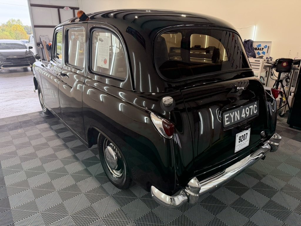 The rear three quarters of the taxi in the detailing shop, black paint reflecting the shop lights, chrome bumper, London plate EYN 497C above a white plate reading 9207.